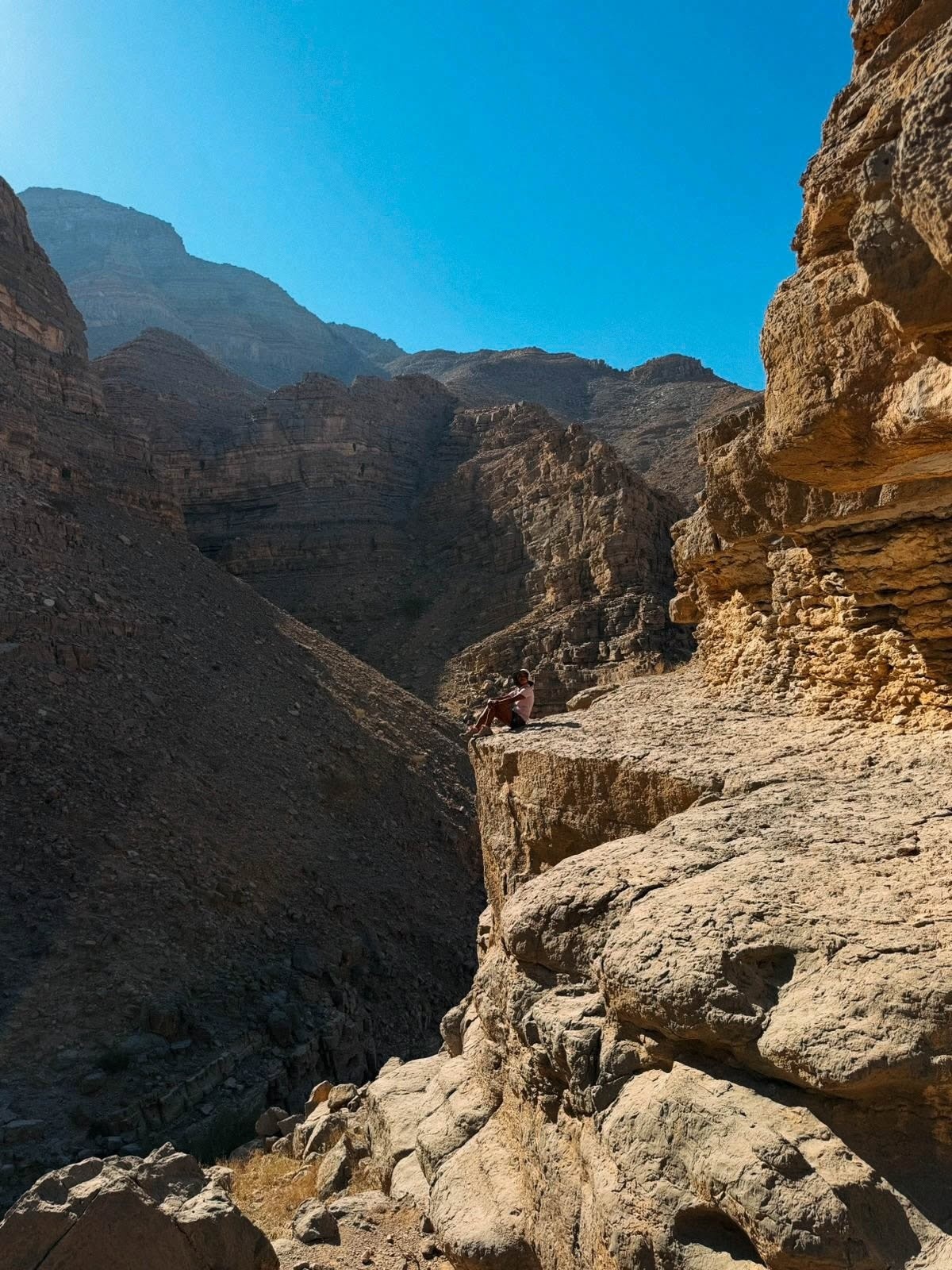 Person sitting on rocky cliff in mountainous desert landscape under clear blue sky.