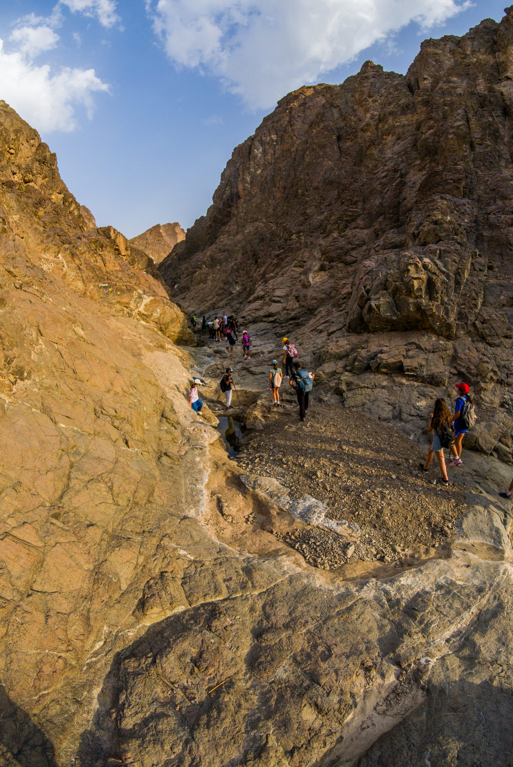 Group hiking through a rocky canyon under a blue sky with clouds.