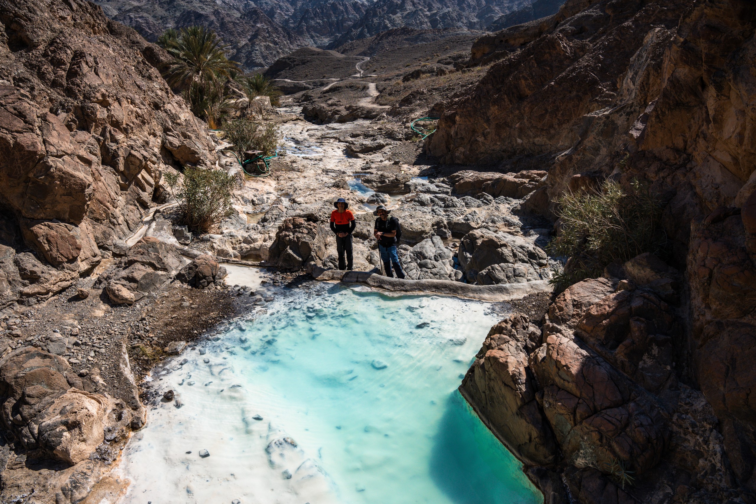 Two people stand near a turquoise pool surrounded by rocky landscape in a canyon.