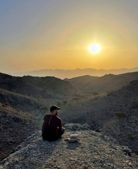 Person sitting on rocky hill at sunset with mountain backdrop.