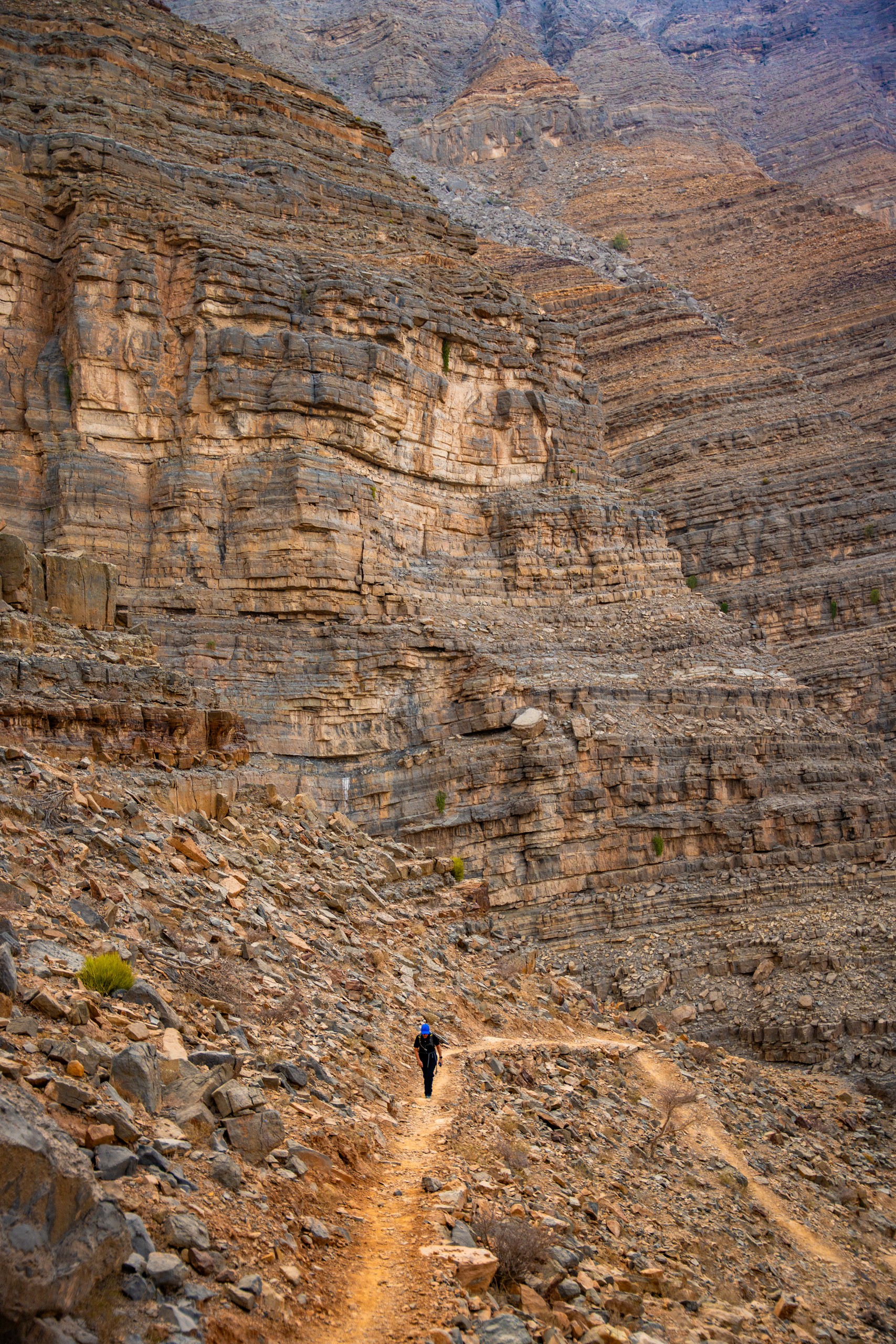 Person hiking on a rocky mountain path with steep, layered cliffs.