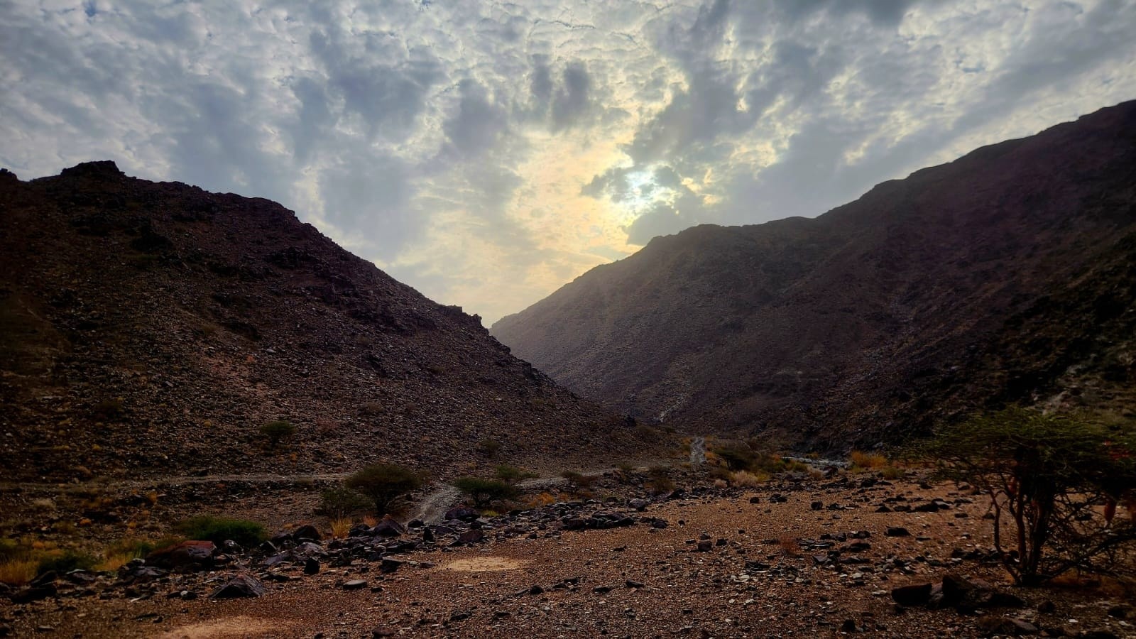 Mountain valley at sunset with cloudy sky and rocky terrain.